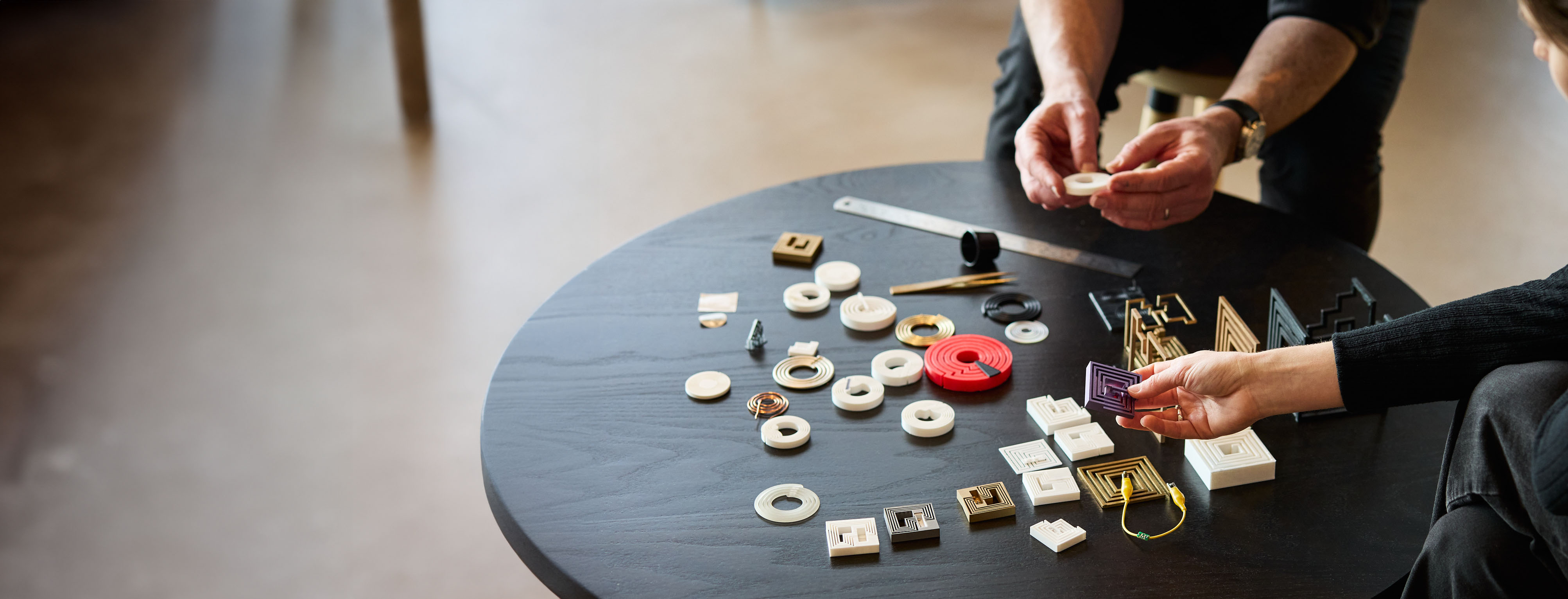 Various NFC antenna prototypes and components arranged on a black round table, with hands working with circular white rings, gold square patterns, and a distinctive red circular component